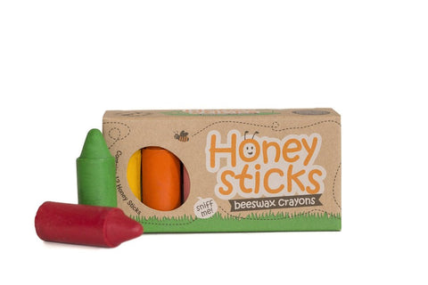 A set of Honeysticks Beeswax Originals Crayons (12 pk) is shown. The box, made of brown cardboard with the Honeysticks brand name and design printed on it, highlights that these non-toxic crayons are handmade in New Zealand. Three crayon colors—green, orange, and red—are visible, with one green and one red crayon placed in front of the box.