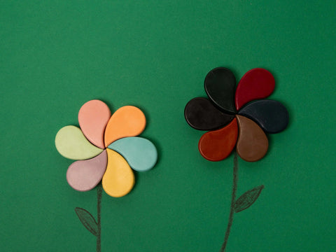 Two flowers are drawn on a green background using Honeysticks Petal Shaped Crayons from Honeysticks, made from 100% pure New Zealand beeswax. The flower on the left features lighter, pastel-colored petals, while the flower on the right showcases darker, more muted-colored petals. Both flowers boast simple stems and leaves that emphasize their non-toxic and safe design.