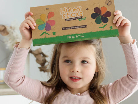 A young girl with light brown hair holds up a box of "Honeysticks Petal Shaped Crayons (12 pk)" over her head. The brown box, adorned with crayon-colored flower designs, reads "12 petal-shaped crayons." Made from 100% pure New Zealand beeswax, the non-toxic and safe crayons make her smile slightly in her pink long-sleeved shirt.