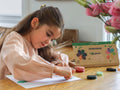 A young girl with long brown hair sits at a wooden table drawing with colorful Honeysticks Petal Shaped Crayons (12 pk), made from 100% pure New Zealand beeswax. She is focused on her work. There is an open box of Honeysticks non-toxic and safe crayons on the table, and a vase with pink flowers in the foreground.