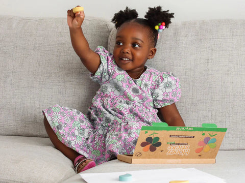 A young girl with two buns in her hair, adorned with colorful beads, is sitting on a gray couch. She wears a pink and gray floral dress and holds up a piece of chalk excitedly. In front of her is an open box of non-toxic and safe Honeysticks Petal Shaped Crayons (12 pk), made from 100% pure New Zealand beeswax.