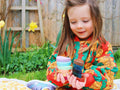 A young child with long hair and a colorful floral jacket sits outdoors, holding a stack of Honeysticks Petal Shaped Crayons (12 pk) made from 100% pure New Zealand beeswax. They are looking down at the non-toxic, safe items with a curious expression. There are green plants and daffodils in the background.