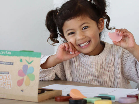 A young girl with pigtails and a white sweater smiles while holding up a pink paint sticker. In front of her is a set of Honeysticks Petal Shaped Crayons (12 pk) alongside drawing papers on the table. A box labeled "100% pure New Zealand beeswax" with a flower illustration, from the brand Honeysticks, is also on the table. The crayons are colorful, non-toxic, and safe for use.