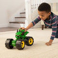 A young child is kneeling on a carpeted floor, playing with a John Deere Monster Treads All-Terrain Tractor, its large black and yellow wheels perfect for all-terrain fun. The background features a staircase and a railing, suggesting the setting is a cozy home interior.