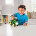 A young child with curly hair is smiling and playing with a green John Deere Roll & Go Flashlight on a white table. The child, wearing a bright blue shirt, is in a room with white walls, shelves stocked with books, and a window in the background. The toy emits engine sounds that delight the child.