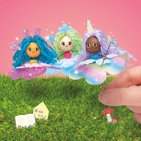 Three whimsical dolls from the Klutz Enchanted Fairy House by KLUTZ, dressed in colorful outfits, stand on grass against a pink background. Each fairy has unique, vibrant hair and wings; one of them is holding a unicorn horn. A hand places a tiny fairy-sized storybook next to a mushroom and another miniature book on the grass.