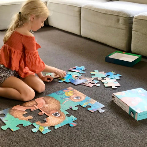 A young girl with blonde hair and an orange off-shoulder top is sitting cross-legged on a carpet. She is assembling the Kuwi's Rowdy Crowd Floor Puzzle - 24 Large Pieces by KUWI THE KIWI, featuring cartoon animal images reminiscent of illustrations from a children's book. Nearby, the open puzzle box rests beside a section of a beige couch.