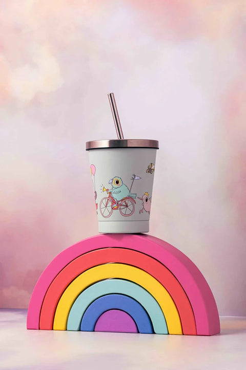 The Kuwi's The Kiwi Stainless Steel Smoothie Cup by KUWI THE KIWI, which is BPA-free and comes with a lid and straw, showcases charming illustrations of a bird riding a bicycle. It is positioned atop a vibrant, stacked rainbow block against a backdrop of softly blended pastel colors.