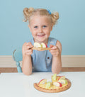 A young girl with blond hair in pigtails is sitting at a table, holding a slice from the Le Toy Van Apple Tart Wooden Playset by LE TOY VAN, crafted from sustainable rubberwood. She is smiling and wearing a sleeveless blue dress. The background features a light blue wall with a white-bordered baseboard.