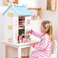 A young child wearing pink and white striped pajamas plays with the Le Toy Van Blue Bird Cottage with Furniture by LE TOY VAN. The pastel-decorated dollhouse is filled with a 37-piece furniture set and dolls. The child sits on a white chair, reaching into the open dollhouse door with a pink towel.