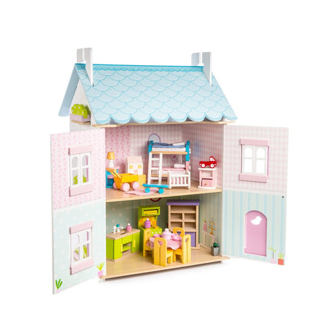 The Le Toy Van Blue Bird Cottage with Furniture by LE TOY VAN is a vibrant wooden dollhouse adorned with blue roof shingles and a pastel pink and green exterior. This charming cottage boasts two stories featuring various rooms, complemented by a comprehensive 37-piece furniture set that includes a kitchen, dining area, bedroom with bunk beds, and nursery. The front doors open to reveal the beautifully detailed interiors.