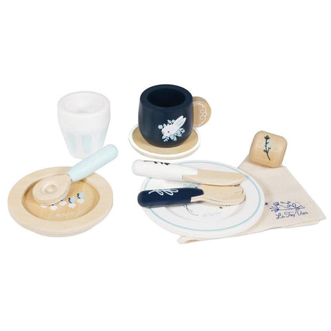 Introducing the *Le Toy Van Cutlery Dining Set by LE TOY VAN, perfect for imaginative role-play. This delightful toy set features wooden cups, saucers, utensils, and a fabric napkin. The set includes a light blue handle on a beige saucer, a blue cup with its matching saucer, and a wooden cutlery set comprising of a knife, fork, and spoon. Completing the ensemble is a charming cloth napkin adorned with blue and white patterns.