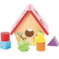 The LE TOY VAN Little Bird House Shape Sorter is a wooden bird house shape sorter with a vibrant red roof designed to develop pre-reading skills. Featuring a central round hole and various shaped cutouts on the roof, this product is crafted from sustainable wood. It includes colorful shapes such as a cylinder, cube, heart, cross, and triangle placed around the house.

