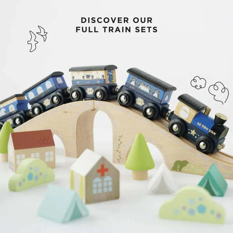 A wooden toy train set, named the Le Toy Van Twilight Train by LE TOY VAN, is shown featuring blue train cars on a curved wooden bridge. Small wooden trees, houses, and tents crafted from sustainable rubberwood are placed around the train. The text "DISCOVER OUR FULL TRAIN SETS" is displayed at the top of the image.