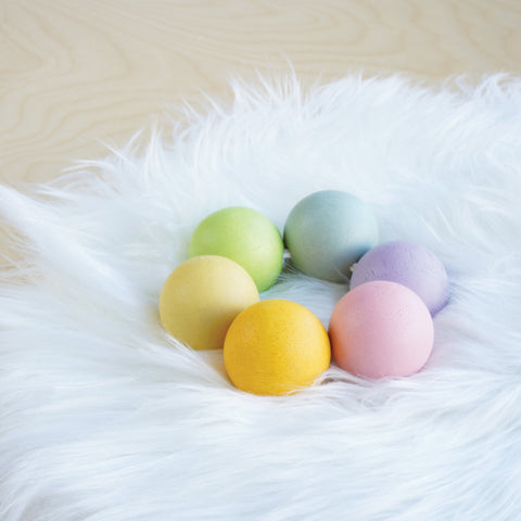 Pastel-colored eggs in shades of yellow, pink, green, blue, and purple are displayed on a fluffy white surface. They are arranged in a semi-circle with Le Toy Van Wooden Beads by LE TOY VAN interspersed. The background features a light wood texture.