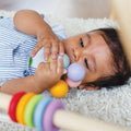 A baby lies on a soft, textured rug, holding and examining a colorful, multi-textured baby-safe toy in their hands. In the foreground, there is the LE TOY VAN Le Toy Van Wooden Beads with rainbow-colored rings made from sustainable rubberwood. The baby is wearing a striped outfit and looks focused on the toy.