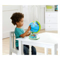 A young child with curly hair sits at a white table, playing with the LeapFrog LeapGlobe Touch by Vtech—an interactive learning globe featuring buttons and educational elements. The room is bright and minimalistic, adorned with white furniture.