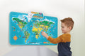 A young boy interacts with the vibrant, wall-mounted Leapfrog Touch & Learn World Map by Leapfrog. This educational toy showcases illustrations of animals, landmarks, and continents, and includes a speaker device on top that provides auditory information. He is smiling and appears engaged in the touch-and-learn activity.