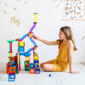 A child in a yellow outfit is sitting on the floor, playing with the Learn & Grow Rainbow Magnetic Tiles Ball Run Pack 88pc, which includes various shapes, tubes, and an exciting magnetic ball run. The wall behind them is decorated with a floral design and a poster featuring butterflies and insects. A sign reading "Play" is also visible on the wall.