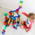 A child with curly hair sits on the floor playing with an 88-piece Learn & Grow Rainbow Magnetic Tiles Ball Run Pack. The set, designed by Learn & Grow, is made of transparent pieces in a variety of colors and includes ramps, loops, and tunnels. The child is wearing a red and white shirt with red shorts.