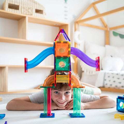 A child lies on the floor, smiling, behind a colorful structure built with the **Learn & Grow Rainbow Magnetic Tiles Ball Run Pack 88pc**. The structure features various shapes, such as squares, triangles, and arches. Shelves and a playhouse frame are visible in the background.