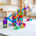 A vibrant and colorful marble run toy set up in a living room with soft toys and a bookshelf in the background. The Learn & Grow Rainbow Magnetic Tiles Ball Run Pack 88pc by Learn & Grow features various transparent cubes and slide pieces in multiple colors, through which marbles can roll.