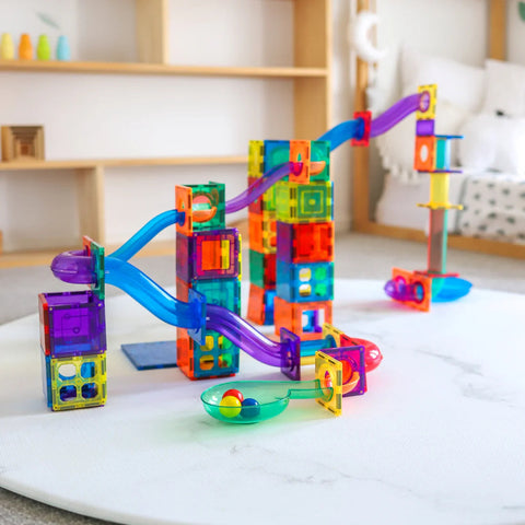 A vibrant and colorful marble run toy set up in a living room with soft toys and a bookshelf in the background. The Learn & Grow Rainbow Magnetic Tiles Ball Run Pack 88pc by Learn & Grow features various transparent cubes and slide pieces in multiple colors, through which marbles can roll.