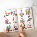 A child's hand pointing to colorful and intricate structures built from the Learn & Grow Rainbow Magnetic Tiles Ball Run Pack 88pc displayed in a book. The book is on a white textured surface and showcases a variety of designs using the multicolored blocks, including an impressive ball run made with an expansion pack of magnetic tiles from Learn & Grow.