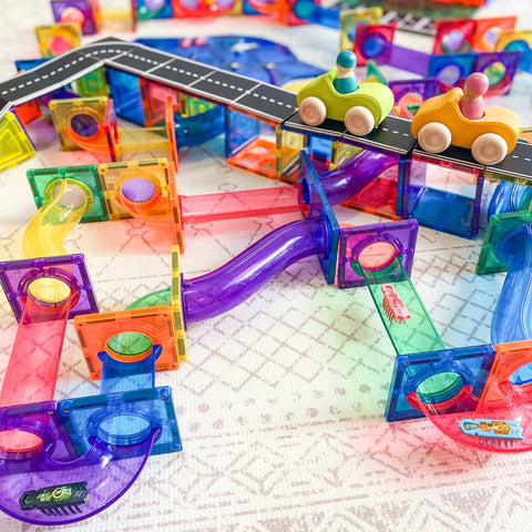 The "Learn & Grow Rainbow Magnetic Tiles Ball Run Pack 88pc" by Learn & Grow is assembled on a patterned surface, showcasing its colorful and transparent plastic pieces. Two toy cars, each featuring a round peg figure, are positioned on a black and white toy road elevated above the magnetic ball run tracks.