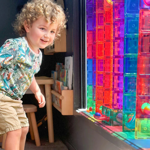 A small child with curly hair, wearing a tropical-themed shirt and beige shorts, is standing next to a window adorned with colorful translucent tiles from the Learn & Grow Rainbow Magnetic Tiles Ball Run Pack 88pc. Sunlight filters through the tiles, casting vibrant hues on the child's fascinated face.