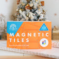 The image shows a brightly colored box labeled "Learn & Grow Rainbow Magnetic Tiles Builders Pack 110pc" from Learn & Grow. The orange and blue design rests on a wrapping paper-covered table with a decorated Christmas tree in the background, ideal for building STEM skills and enhancing both fine and gross motor skills.