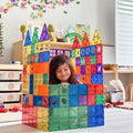 A young child with brown hair sits joyfully inside a vibrant castle made of interlocking pieces from the Learn & Grow Rainbow Magnetic Tiles Builders Pack 110pc, enhancing their fine and gross motor skills. The castle, boasting multiple pointed towers, stands in a playroom adorned with shelves, toys, and butterfly decorations on the wall.