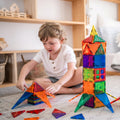 A young child with short hair and a white shirt is sitting on the floor, playing with the Learn & Grow Rainbow Magnetic Tiles Builders Pack 110pc. They have constructed a tall tower and are building another structure, developing fine and gross motor skills. Shelves with organized baskets and toys are in the background.