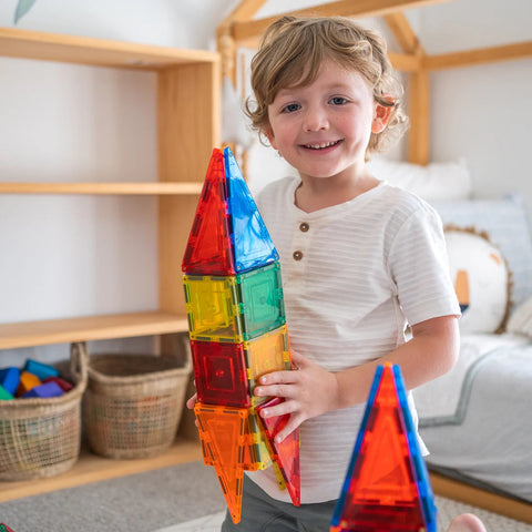 A young child with shoulder-length hair smiles while holding a large, colorful rocket-shaped structure made from the Learn & Grow Rainbow Magnetic Tiles Builders Pack 110pc in a playroom. The room, designed to encourage STEM skills, features wooden shelves, baskets, and additional toys from Learn & Grow that foster fine and gross motor skills.