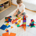 A child sits on a patterned rug, playing with the Learn & Grow Rainbow Magnetic Tiles Builders Pack 110pc. They’ve built several structures, including two rocket-like shapes, and are in the process of stacking more tiles. Shelves and baskets filled with toys are visible in the background, fostering their STEM skills and motor development.