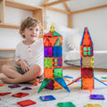 A young child sits on the floor, smiling while playing with colorful **Learn & Grow Rainbow Magnetic Tiles Builders Pack 110pc**. Two large structures resembling rockets are constructed using the tiles, and various other tiles are scattered around the carpet, aiding in developing fine and gross motor skills. A wooden shelf is partially visible in the background.