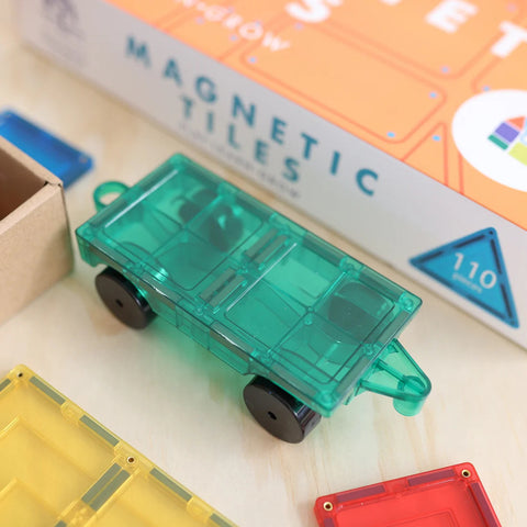 A green, transparent, magnetic toy train rests on a wooden surface. Surrounding it are colorful Learn & Grow Rainbow Magnetic Tiles in red, yellow, and blue. A partially visible box of Learn & Grow Rainbow Magnetic Tiles Builders Pack 110pc in the background indicates the 110-piece set designed to enhance fine and gross motor skills.