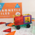 A small toy made of the Learn & Grow Rainbow Magnetic Tiles Builders Pack 110pc is assembled in the shape of a vehicle with wheels, placed on a play mat. Several loose pieces are scattered around, perfect for developing STEM skills. In the background, a partially visible orange box labeled "Learn & Grow" is seen.