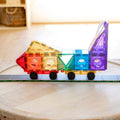 A vibrant toy train crafted using the Learn & Grow Rainbow Magnetic Tiles Car Base Pack 2pc is positioned on a small, green toy road. The train is made up of triangular and square pieces in shades of red, yellow, green, blue, and purple with black wheels. This captivating construction set aids children in developing STEM skills against a backdrop of wooden flooring and shelves.