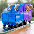 A toy truck crafted from translucent blue and purple plastic blocks with rubber wheels features two beige stuffed mice in the front seats and one in the back, driving on a play mat. The background is blurred greenery. This set from Learn & Grow can be expanded with the Learn & Grow Rainbow Magnetic Tiles Car Expansion Pack 28pc.