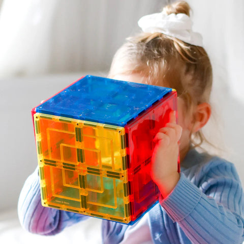 A young child in a blue sweater is holding a vibrant, transparent cube built from interlocking Learn & Grow Rainbow Magnetic Tiles Large Square Pack 8pc set in red, yellow, and blue. With a curious expression, she looks through one side of the cube. A white bow is in her hair, hinting at endless building possibilities offered by Learn & Grow.