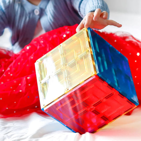 A child in a blue sweater and red skirt is playing with the Learn & Grow Rainbow Magnetic Tiles Large Square Pack 8pc set, creating a large translucent cube made of different colors, including yellow, red, and blue. The child is reaching out to touch the cube, which is placed on a white surface, exploring endless building possibilities from Learn & Grow.