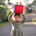 A young girl wearing a leopard print dress holds a red and blue tile from the Learn & Grow Rainbow Magnetic Tiles Large Square Pack 8pc set with two small dolls on top above her head. The background is blurry, suggesting an outdoor setting with greenery and a clear sky, perfect for building possibilities.