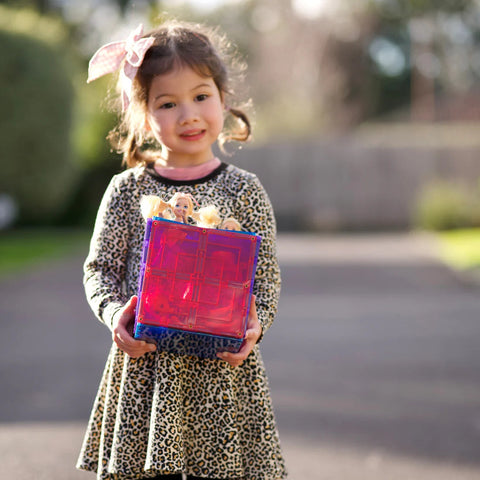 A young girl with a light pink bow in her hair wears a leopard print dress and smiles while holding a large, semi-transparent pink magnetic tile from the Learn & Grow Rainbow Magnetic Tiles Large Square Pack 8pc set. Some small doll heads peek out from the top of the tile block. She stands on a paved area outdoors, embracing endless building possibilities with her Learn & Grow tiles.