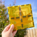 A hand holding a yellow transparent square case with a pressed, brownish-yellow leaf inside is showcased against a dreamy backdrop of leafy greenery and part of a wooden fence under a blue sky, reminiscent of the endless building possibilities offered by the Learn & Grow Rainbow Magnetic Tiles Large Square Pack 8pc set from Learn & Grow.