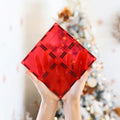 Two hands are holding up a red, diamond-shaped object made of interlocking translucent pieces from the Learn & Grow Rainbow Magnetic Tiles Large Square Pack 8pc set. The background features a decorated Christmas tree with blurred lights and ornaments, showcasing the building possibilities of this innovative set by Learn & Grow.