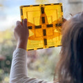 A person with long hair, viewed from behind, holds up a yellow translucent tile from the Learn & Grow Rainbow Magnetic Tiles Large Square Pack 8pc set towards the light. The panel contains a pressed leaf, revealing its intricate veins and structure. The background is softly blurred with hints of foliage and light, suggesting endless building possibilities with Learn & Grow.