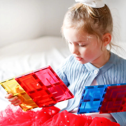 A young child with blonde hair, wearing a blue cardigan and a red skirt, is sitting and holding colorful transparent magnetic tiles from the Learn & Grow Rainbow Magnetic Tiles Large Square Pack 8pc set by Learn & Grow. The tiles are red, yellow, and blue. The child appears to be focused on building possibilities in a well-lit indoor setting.