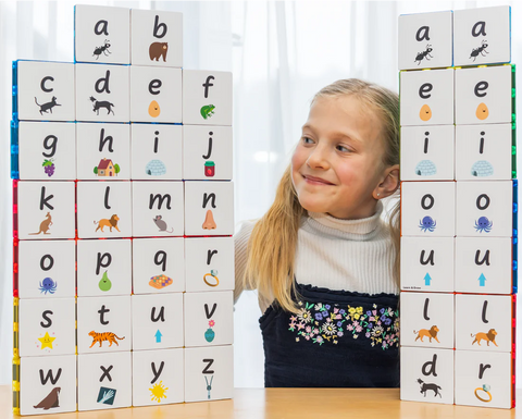 A young girl smiles while looking at two stacks of Learn & Grow Rainbow Magnetic Tiles Toppers - Alphabet Lower Case Pack 40pc on a table. The tiles, an educational tool, feature lowercase alphabet letters along with images representing words that start with each letter, such as animals and objects. She is seated behind the stacks in a brightly lit room.