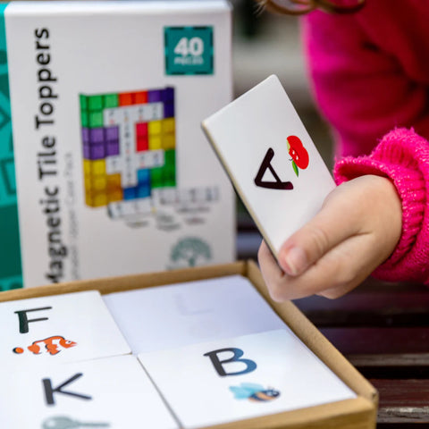 A child in a bright pink sleeve is holding a Learn & Grow Rainbow Magnetic Tiles Topper featuring an uppercase letter A with an apple illustration. In the background, there are more magnetic letter tiles and a box labeled "Learn & Grow Rainbow Magnetic Tiles Toppers - Alphabet Upper Case Pack 40pc," perfect for early literacy instruction.